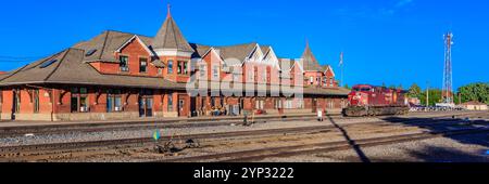 Un treno sta entrando in una stazione ferroviaria con un grande edificio rosso sullo sfondo Foto Stock