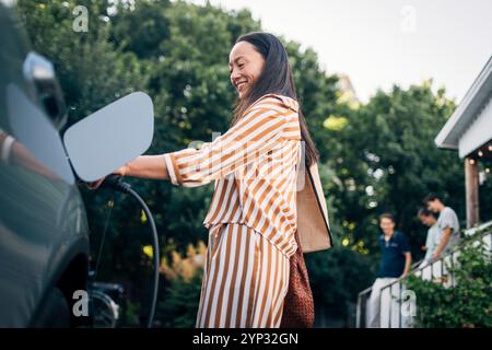 Visuale ad angolo basso della donna sorridente che carica l'auto con la spina elettrica nel cortile posteriore Foto Stock
