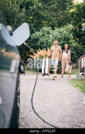 Coppia felice che trasporta bagagli mentre cammina sul sentiero verso la ricarica dell'auto nel cortile posteriore Foto Stock