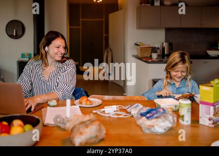 Mamma sorridente con un portatile che guarda il figlio spalmare il burro sul pane a casa Foto Stock