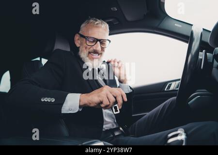 Un uomo d'affari maturo regola la cintura di sicurezza dell'auto mentre si prepara alla guida, mostrando i concetti di viaggio e di lavoro in abbigliamento formale Foto Stock