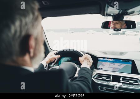 Uomo d'affari maturo che guida un'auto in città, indossa una tuta formale, incentrata sulla navigazione e sui dettagli del viaggio Foto Stock