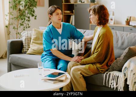 Assistente in uniforme blu seduta con una donna anziana che indossa gli occhiali, tiene la mano e offre supporto emotivo in un soggiorno accogliente con arredi caldi Foto Stock