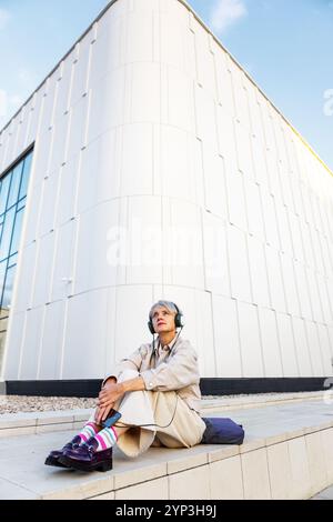 Bella donna di mezza età alla moda con capelli corti e cuffie in abiti beige seduti all'aperto in città, ascoltando musica, podcast o audiolibri Foto Stock