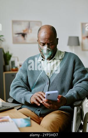 Ritratto di un uomo anziano seduto in sedia a rotelle, che indossa una maschera di ossigeno mentre utilizza lo smartphone, guardando leggermente verso il basso Foto Stock