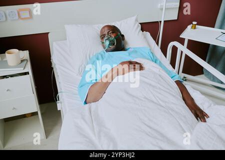 Paziente afroamericano sdraiato nel letto d'ospedale indossando maschera di ossigeno e abito blu, situato in una stanza medica con biancheria da letto bianca, e collegato a gocciolamento endovenoso Foto Stock