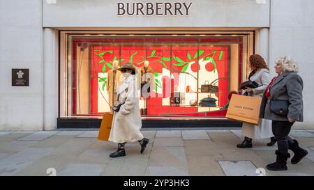 Londra, Regno Unito. 28 novembre 2024. Gli acquirenti che portano le borse Louis Vuitton passano le decorazioni natalizie nelle vetrine del negozio Burberry a New Bond Street nel West End. New Bond Street è stata recentemente votata la terza via dello shopping più costosa al mondo con via Montenapoleone di Milano che sorpassa la Upper 5th Avenue di New York per rivendicare il primo posto. Crediti: Stephen Chung / Alamy Live News Foto Stock