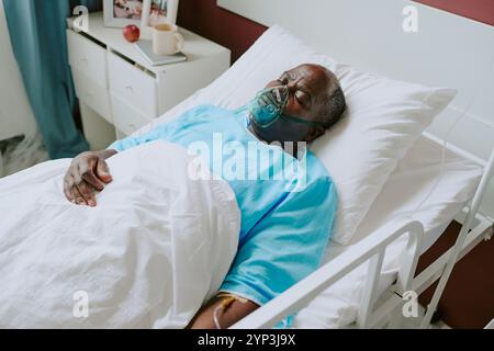 Elderly man lying in hospital bed, wearing blue gown and using oxygen mask, open curtains and bedside table visible with personal items in hospital room Foto Stock