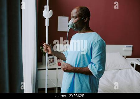 Uomo anziano in piedi accanto alla finestra in stanza d'ospedale indossando maschera di ossigeno e abito, contemplando il pensiero mentre tenendo IV stand, catturato in ambiente medico Foto Stock