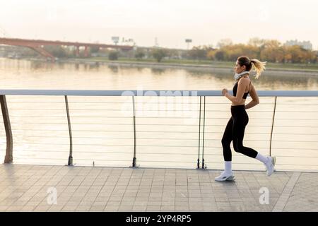 Un determinato corridore procede con sicurezza lungo un sentiero lungo il fiume, abbracciando l'aria frizzante del mattino, con lo skyline della città e gli alberi autunnali che offrono un'immagine Foto Stock