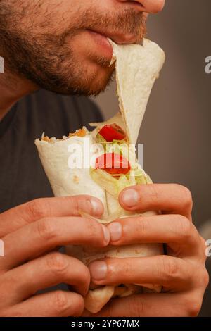 Un uomo con una piccola barba morde in un rotolo di pita pieno di carne, verdure fresche e insalata. La foto, scattata a metà movimento, enfatizza il deliciou Foto Stock