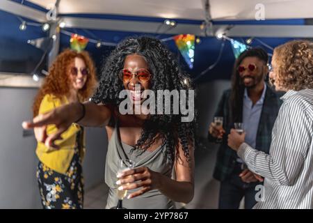 Donna africana che indossa un elegante abito da notte e occhiali da sole puntando avanti a ballare con gli amici durante la festa serale in terrazza Foto Stock