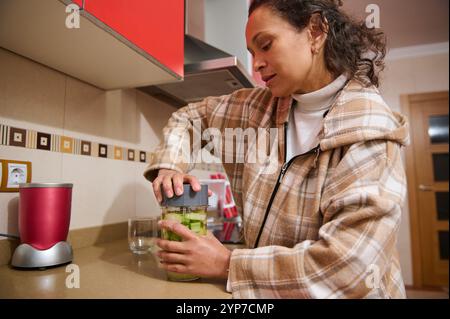 Una donna con una giacca a quadri prepara un frullato verde sano in una cucina moderna. Utilizza un frullatore per mescolare ingredienti freschi, promuovendo una vita sana Foto Stock