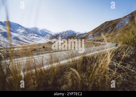 La strada tortuosa conduce attraverso un paesaggio montano erboso e innevato sotto un cielo blu, passo Lindis, nuova Zelanda, Oceania Foto Stock