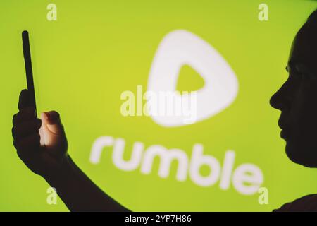 In questa foto, la silhouette di una donna contiene uno smartphone con il logo Rumble sullo sfondo Foto Stock