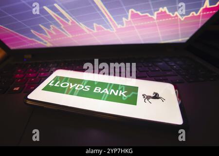 In questa immagine, il logo della Lloyds Bank è visualizzato sullo schermo di uno smartphone, con una rappresentazione grafica del mercato azionario nel backgrou Foto Stock