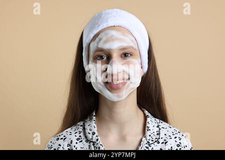 Adolescente con schiuma detergente sul viso su sfondo beige. Trattamento dell'acne Foto Stock