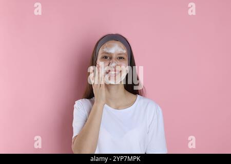 Adolescente con schiuma detergente sul viso su sfondo rosa. Trattamento dell'acne Foto Stock