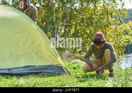 Ragazzo accovacciato sull'erba mentre aiuta suo padre con la tenda lanciante sulla collina, cerca di legare le linee dei Guy Foto Stock