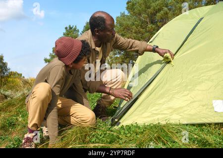 Un colpo di scena basso di padre afroamericano e suo figlio che accovacciano mentre l'uomo spiega come legare le linee di tenda dei Guy Foto Stock