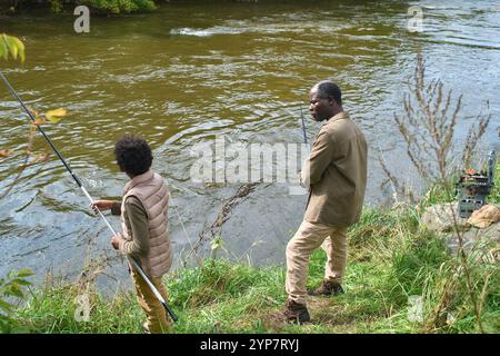 Colpo ad angolo alto di padre e figlio afroamericani che tengono spinnate interagendo mentre pescano sotto cielo aperto Foto Stock
