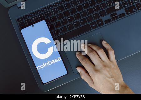 In questa immagine, il logo Coinbase viene visualizzato sullo schermo di uno smartphone accanto a una persona che utilizza un computer portatile Foto Stock