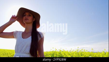 Donna felice che indossa un cappello e cammina nel campo della senape in una giornata di sole Foto Stock