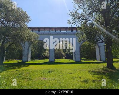 Un grande arco sorregge un sentiero sopra, circondato da una vegetazione vivace e ombreggiato da grandi alberi in una giornata di sole. Foto Stock