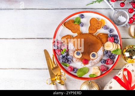 Divertenti e carine frittelle di renna natalizie su un piatto rosso bianco decorato con bacche fresche, frutta e zucchero in polvere, colazione di Natale per bambini Foto Stock