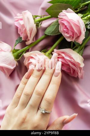 Mano di donna con manicure rosa alla moda e anello di fidanzamento disteso vicino a un bouquet di rose rosa su tessuto di seta Foto Stock