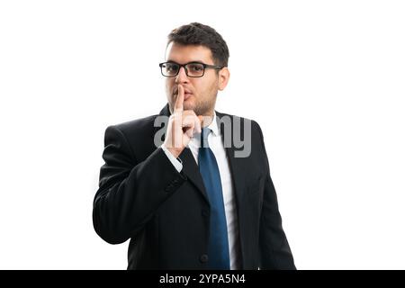 Uomo d'affari adulto dall'espressione seria che indossa un abito casual elegante e occhiali che coprono la bocca con l'indice che rende isolato il gesto segreto dello shush Foto Stock