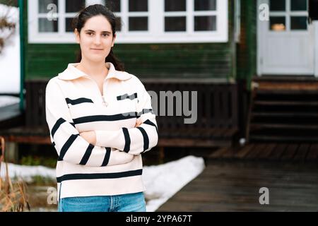 una giovane donna con un maglione a righe si erge davanti a una casa di legno Foto Stock