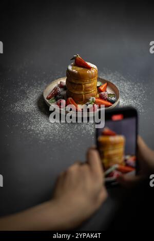 Persona che fotografa un piatto di soffici pancake giapponesi con frutti di bosco freschi, zucchero a velo e sciroppo utilizzando uno smartphone Foto Stock