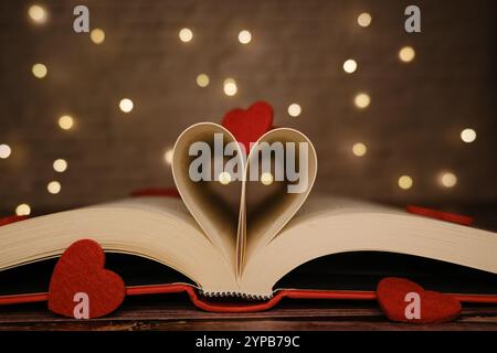 Libro aperto con pagine a forma di cuore, decorate con cuori di feltro rosso e luci bokeh Foto Stock