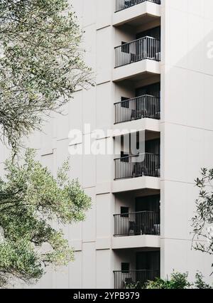 Un moderno edificio di appartamenti con una facciata minimalista che presenta balconi multipli con ringhiere in metallo nero. Circondata da alberi verdi, la struttura Foto Stock