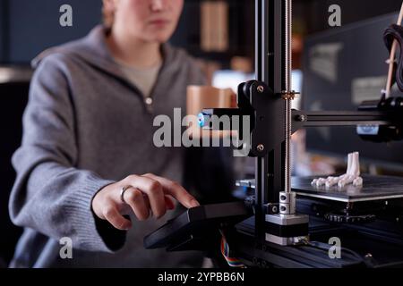 Persona che utilizza una stampante 3D in uno spazio di lavoro moderno, concentrandosi sul funzionamento della macchina con le mani sul pannello di controllo, sfondo sfocato che mostra l'ambiente d'ufficio Foto Stock