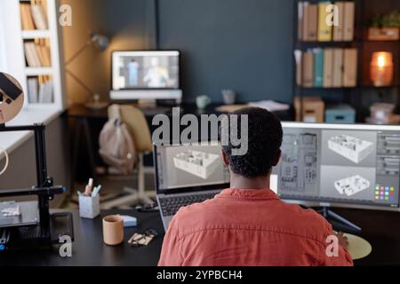 Persona che lavora a un progetto di progettazione 3D con due monitor per computer e una stampante 3D in un ufficio domestico con scaffali e scrivania Foto Stock