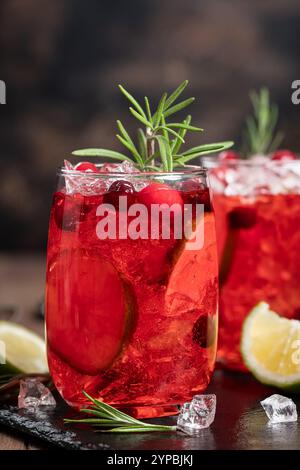 Cocktail di mirtilli rossi con fette di lime e ghiaccio tritato guarnito con rosmarino su tavola di ardesia nera con fondo in legno Foto Stock