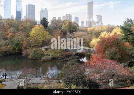 Central Park presenta il suo splendido fogliame autunnale con rossi e oro vivaci. Un affascinante ponte si erge su un tranquillo stagno, incorniciato dallo skyline della città Foto Stock