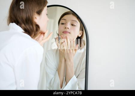 Inquadratura a spalla di una giovane donna attraente che massaggia il viso mentre guarda allo specchio Foto Stock