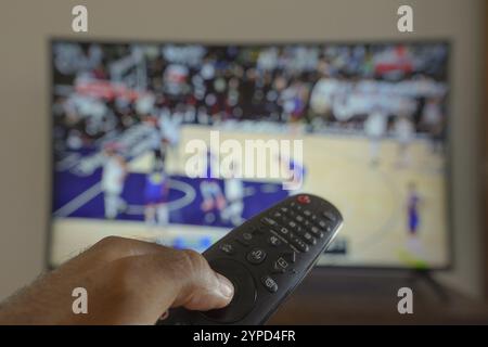 11 maggio 2024, Brasile. In questa illustrazione fotografica, una mano che tiene un telecomando TV davanti alla a che trasmette una partita di basket in TV Foto Stock