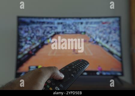 11 maggio 2024, Brasile. In questa illustrazione fotografica, una mano che tiene un telecomando TV davanti alla a che trasmette una partita di tennis sulla TV Foto Stock