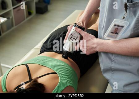 Fisioterapista che tratta il paziente utilizzando un dispositivo specializzato sul lettino portapaziente. Il paziente è adagiato allo stomaco mentre un professionista tiene e utilizza l'apparecchiatura per scopi terapeutici Foto Stock