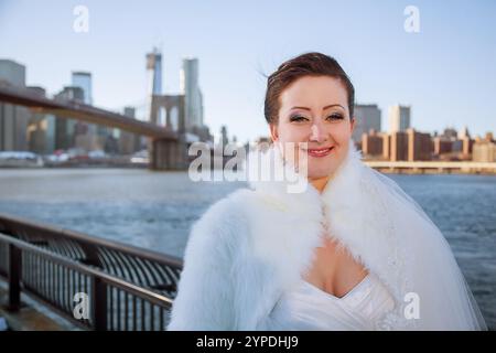 La sposa sorridente si erge sull'acqua, vestita di abito bianco e soffice mantello, con lo splendido skyline di New York alle sue spalle in un luminoso giorno d'inverno Foto Stock