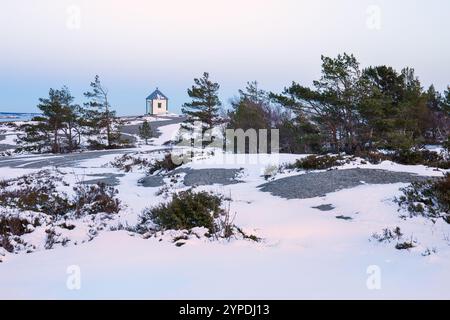 Piccola casa bianca con un tetto blu circondata da terreno innevato e alberi sparsi sotto un pallido cielo invernale. Idilliaco scenario rurale con un tocco minimalista, S. Foto Stock