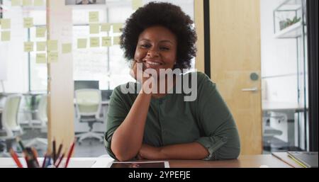 Ritratto di una donna d'affari afroamericana seduta alla scrivania appoggiata alla mano e sorridente in ufficio Foto Stock
