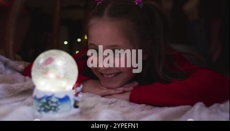 Ragazza caucasica sorridente e guardando il globo di neve mentre si sdraia sotto il forte coperta durante il natale a. Foto Stock