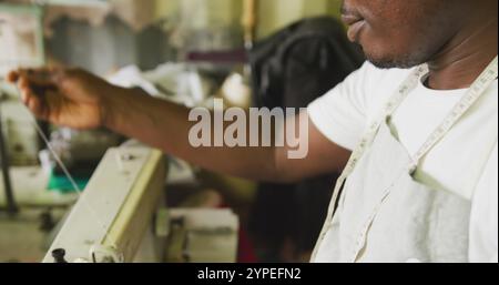 Vista laterale di un sarto africano in un'officina cittadina, seduto accanto a una macchina da cucire e tagliando Foto Stock