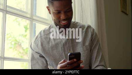 Vista frontale di un giovane uomo nero che utilizza lo smartphone e si siede sul davanzale della finestra in una casa confortevole Foto Stock