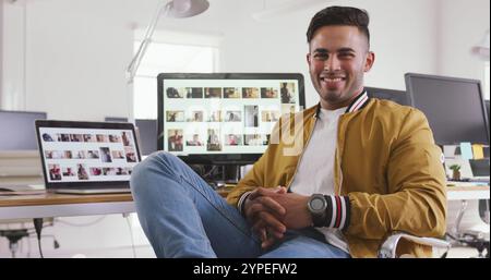 Ritratto di un uomo birazziale felice che lavora in un ufficio creativo, seduto accanto a una scrivania con computer, bagno Foto Stock